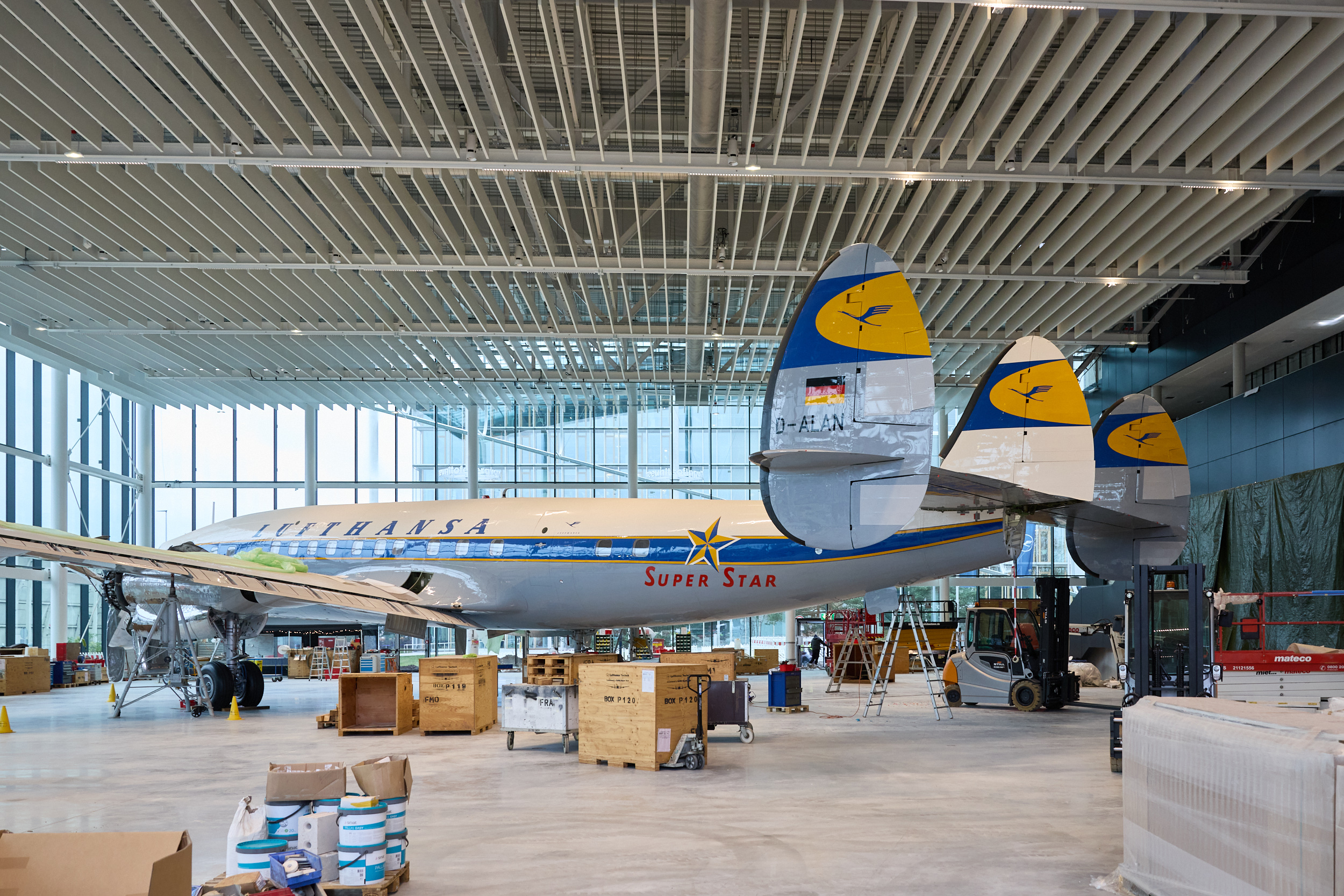 Ein restauriertes historisches Lufthansa-Propellerflugzeug in einer hellen Museumshalle, mit sichtbaren Leitwerken sowie Holzkisten, Leitern und Wartungsausrüstung, die während Aufbau oder Restaurierung darum angeordnet sind.