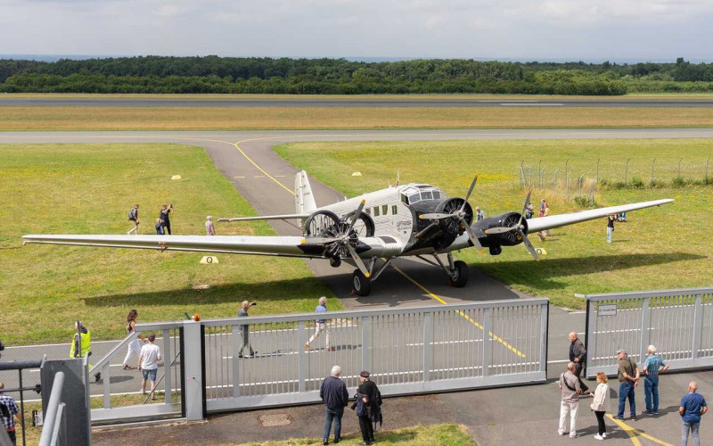 Historische Ju-52 auf einem Rollweg eines Flugplatzes, von hinter einem Zaun aus betrachtet, während Besucher in der Nähe stehen und entlang der Grasflächen neben der Start- und Landebahn gehen.