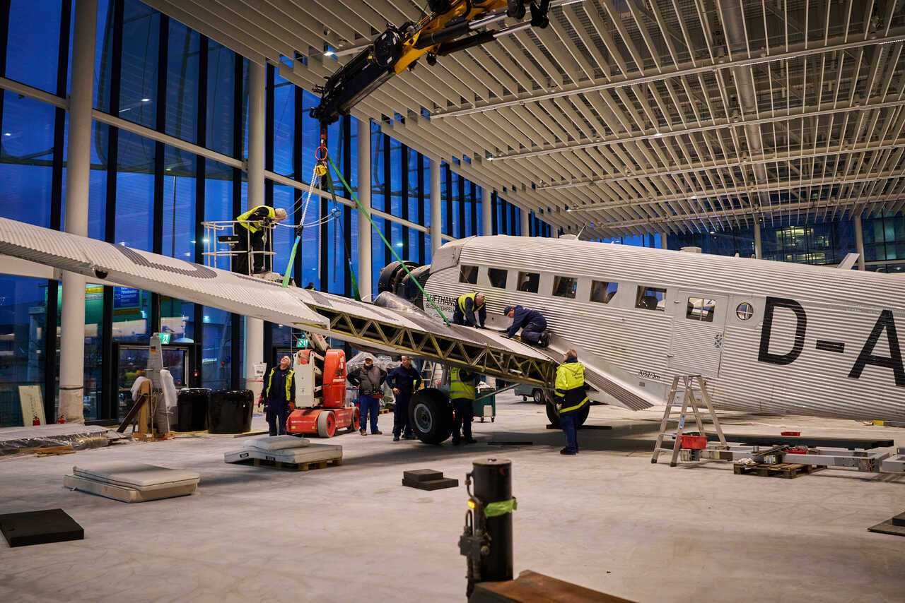 Historisches Lufthansa-Propellerflugzeug in einer großen, verglasten Museumshalle, während mehrere Arbeiter mit einem Kran und Hebegurten den Flügel bei der Montage positionieren; Werkzeuge und Ausrüstung sind um das Flugzeug herum angeordnet.