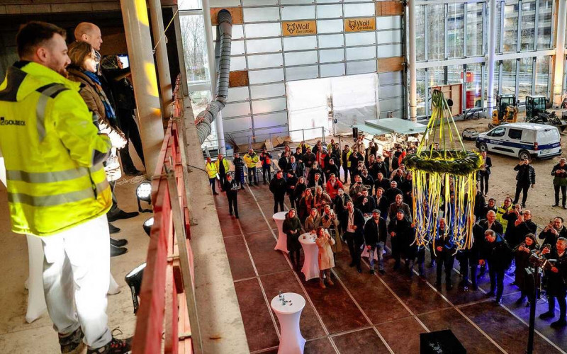 Richtfest in einer Museumshalle: Redner stehen auf einer oberen Ebene und sprechen zu den Gästen darunter, während ein aufgehängter Richtkranz mit bunten Bändern über der versammelten Menge hängt.